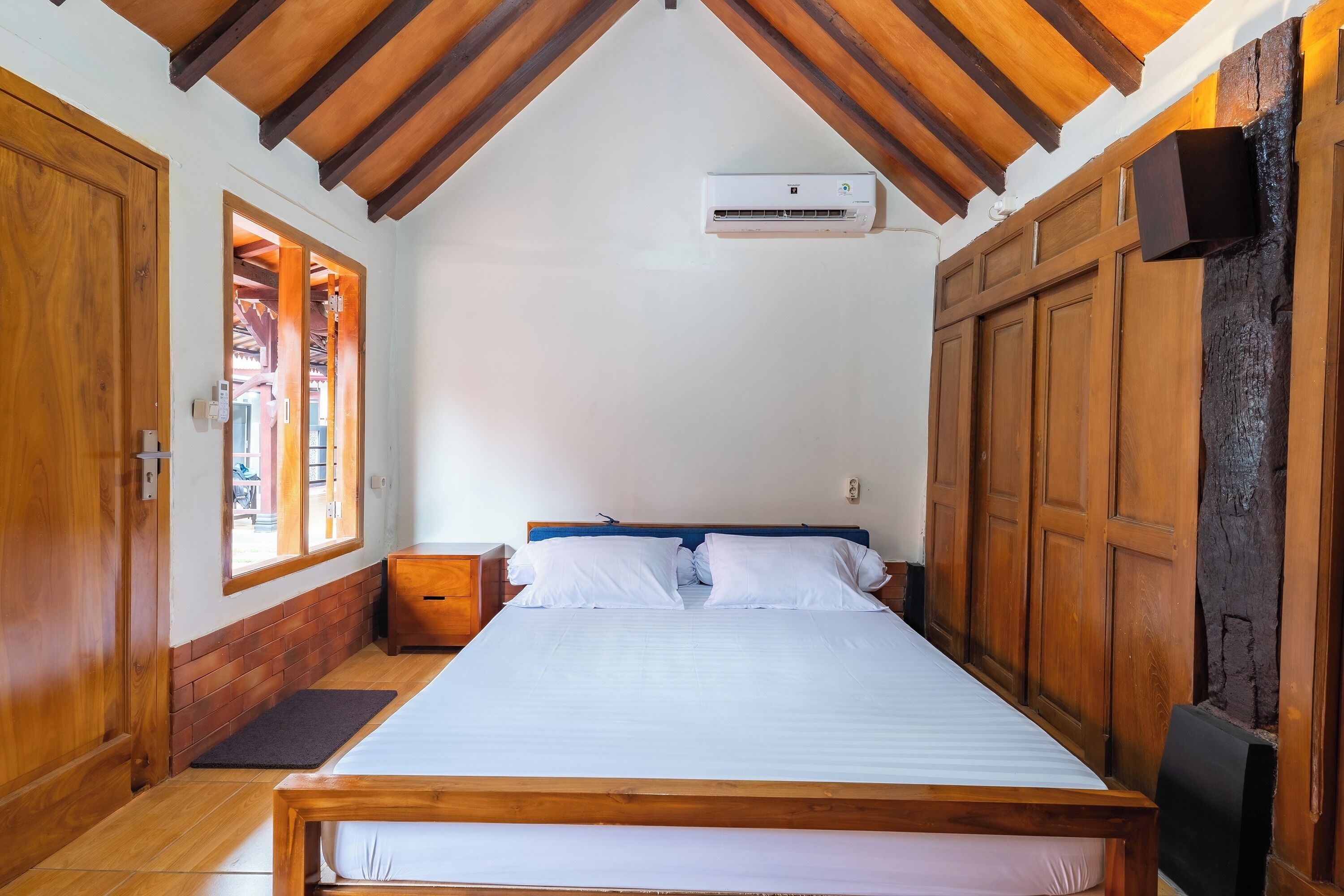 A bedroom with a wooden ceiling, a bed with white bedding, a nightstand, and a wall-mounted air conditioning unit.