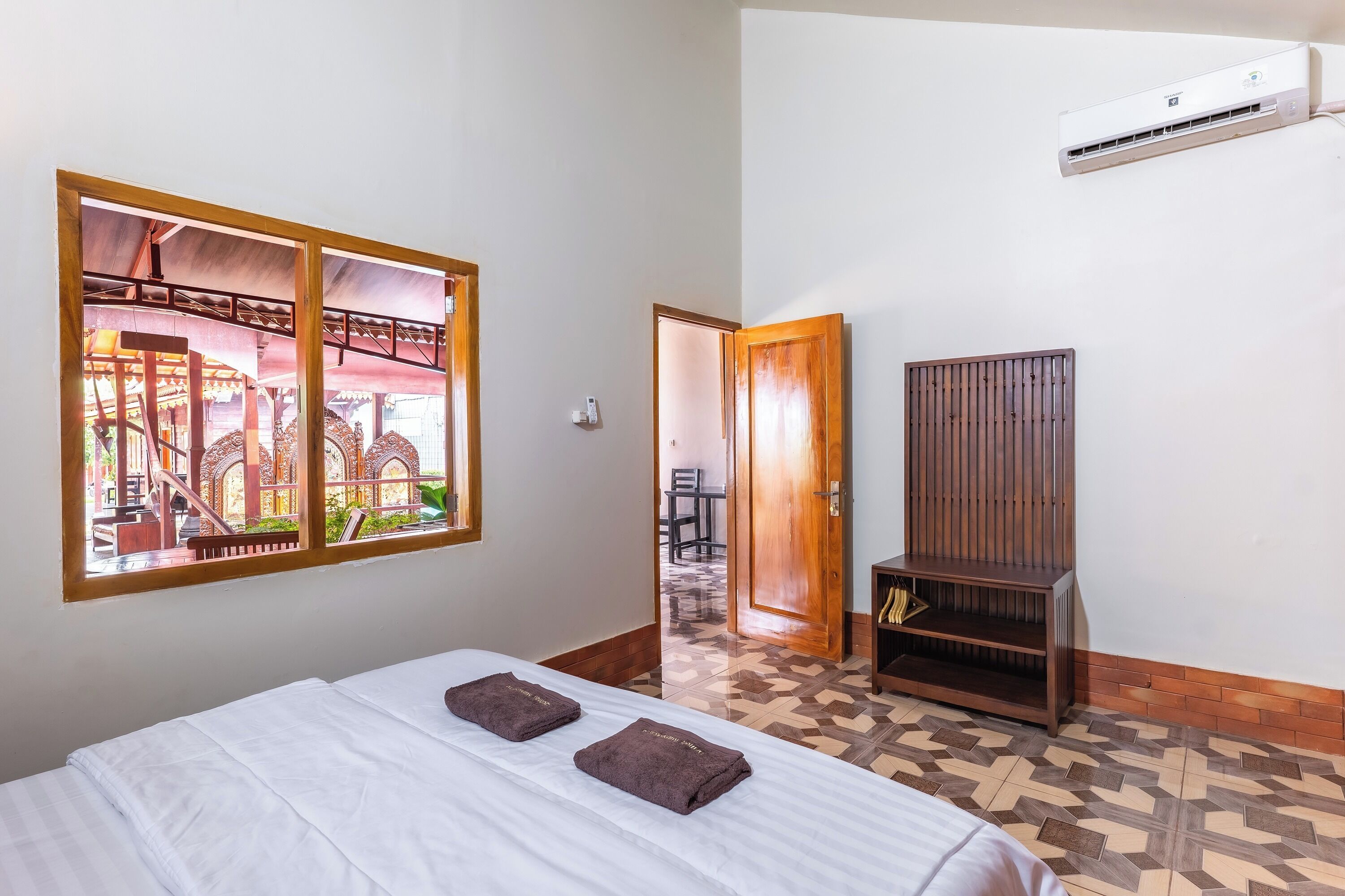 A hotel room with a bed, a wooden wardrobe, a window with a view of a building, and an air conditioning unit.