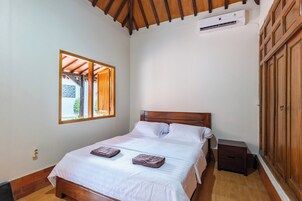 Un dormitorio con cama de madera, ropa de cama blanca, una mesita de noche, un equipo de aire acondicionado y una ventana con vistas.