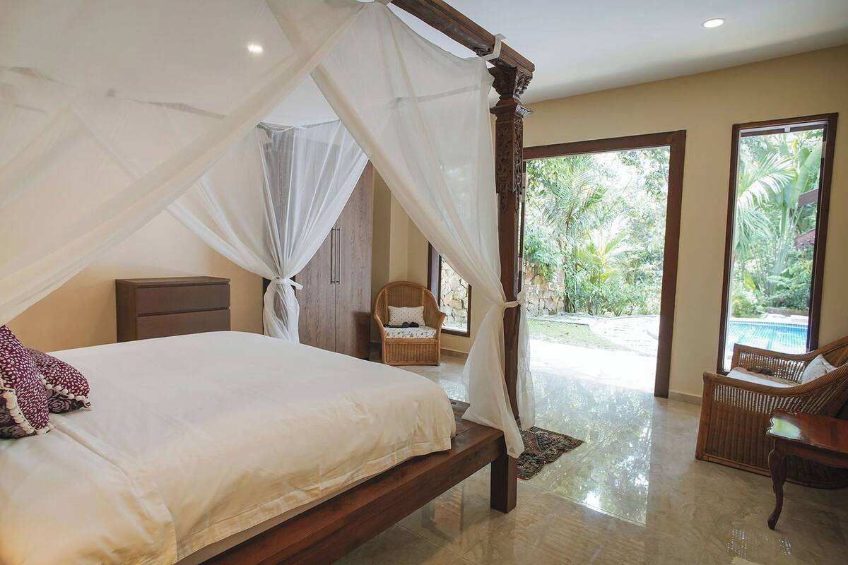 Una cama con dosel, una silla de mimbre y vistas a una piscina.