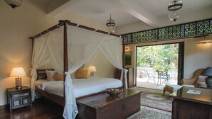 A four-poster bed with a canopy, a wooden chest of drawers, a desk with a lamp, and a view of the garden through a glass door.