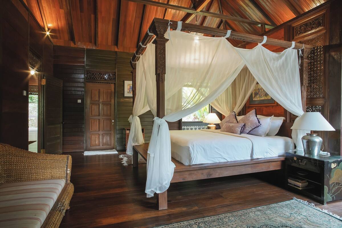 Una cama con dosel de madera, un mesita de noche con lámpara y un sofá de mimbre en una habitación rústica con pisos y paredes de madera.