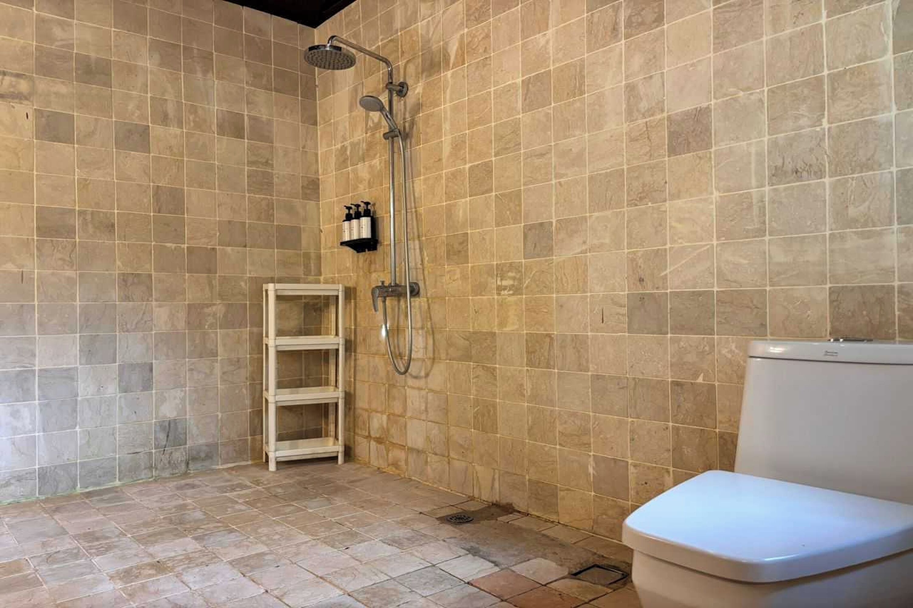A bathroom with beige tiles, a shower, a toilet, and a shelf unit.