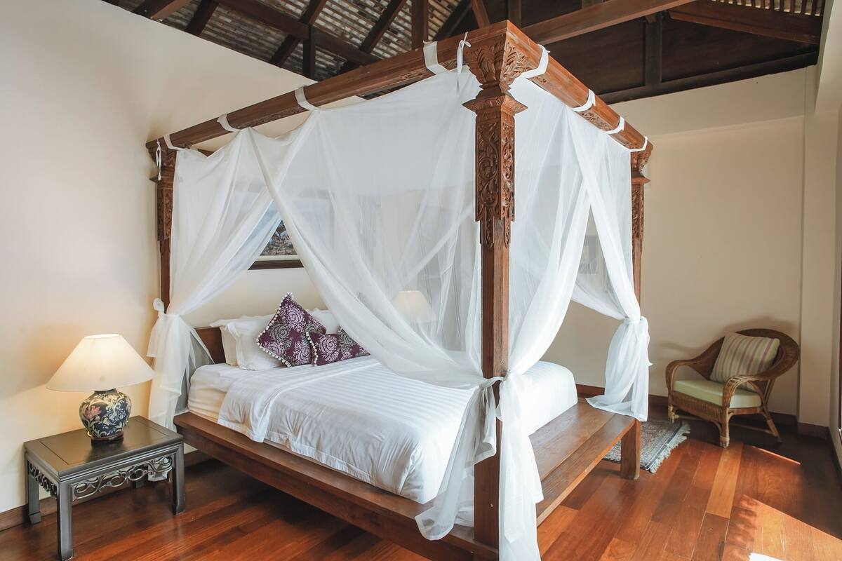Una cama con dosel y cuatro postes, una mesita de noche con lámpara, y una silla en una habitación con piso de madera y techo inclinado.