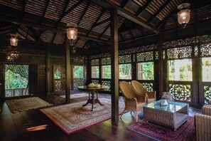 A room with a wooden ceiling, large windows with lattice designs, wicker furniture, and a glass coffee table. - Kebun Rimba Janda Baik (Janda Baik)