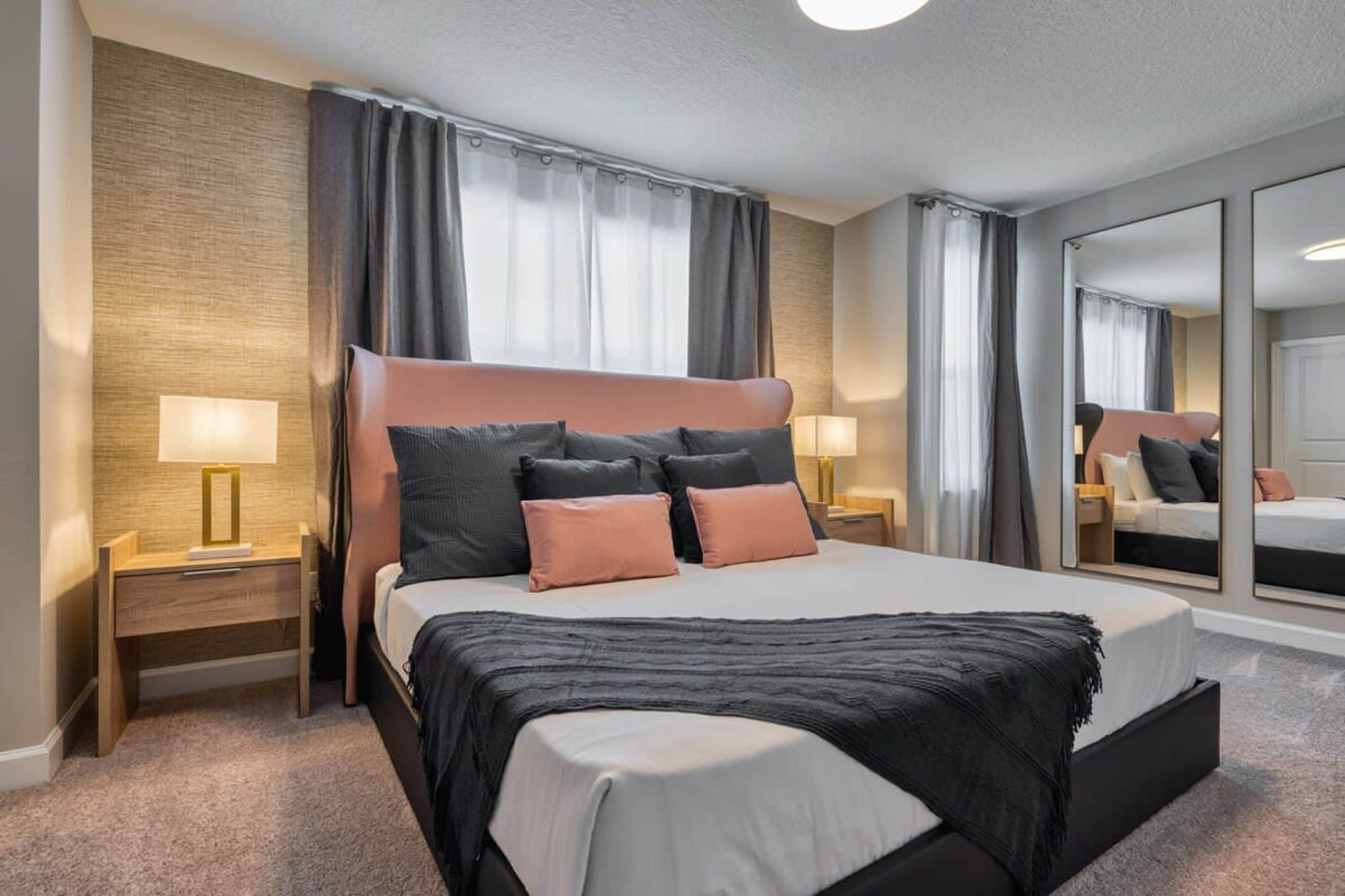 Una moderna camera d'hotel con un letto grande, due comodini con lampade, un ampio specchio e un pavimento rivestito di moquette.