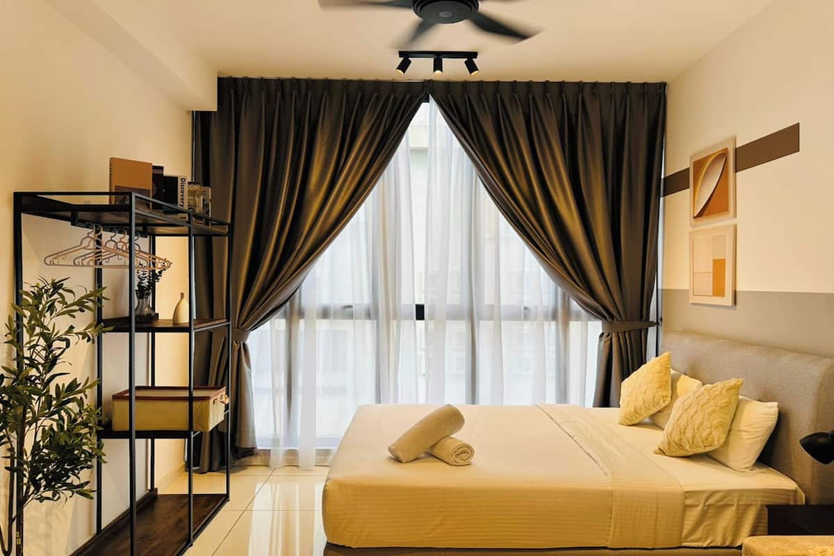 Une chambre d’hôtel avec un lit, un ventilateur de plafond, une fenêtre avec des rideaux, une plante et une étagère avec quelques objets.