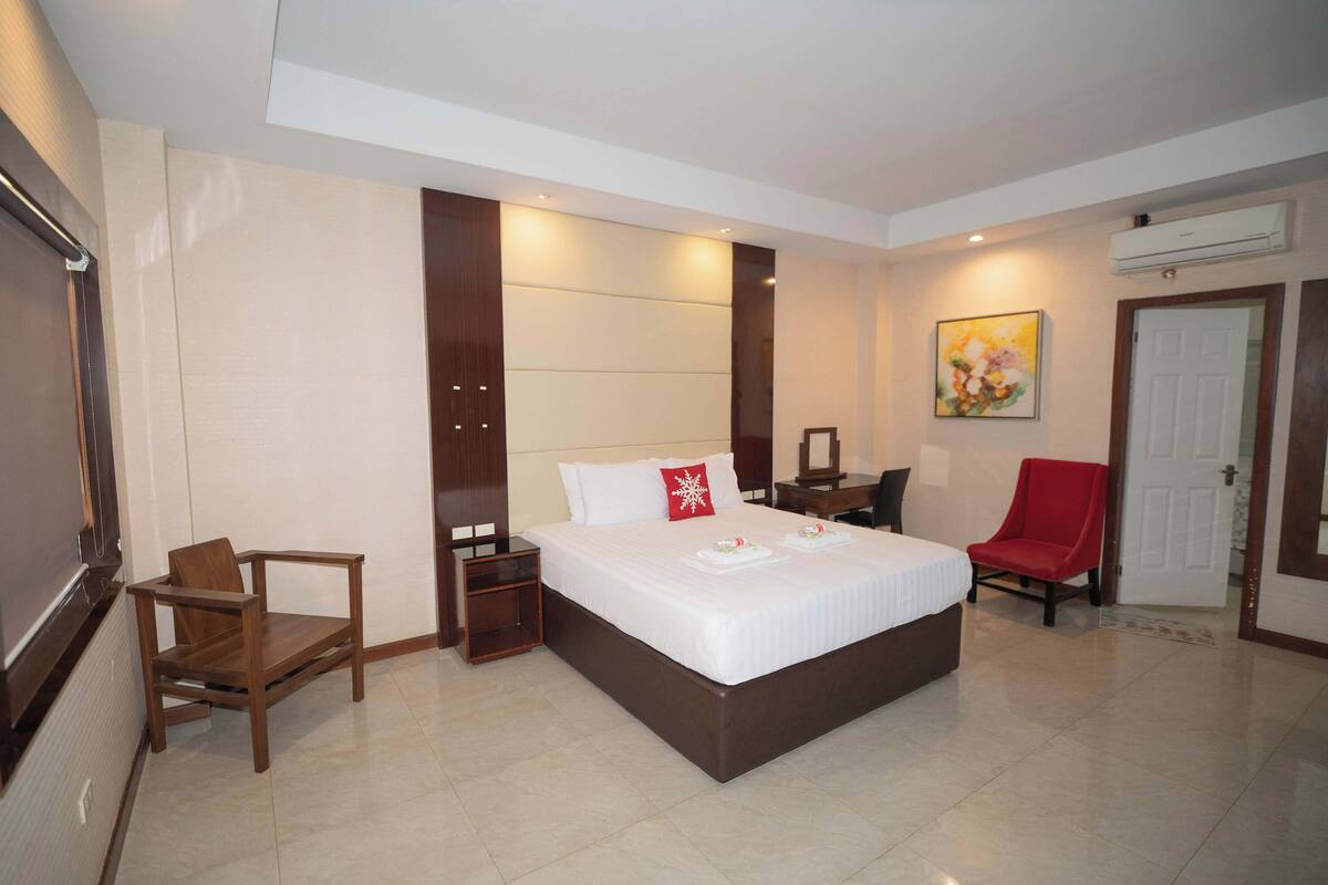 A hotel room with a large bed, a wooden chair, a desk, and an air conditioning unit.