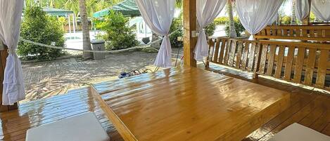 Una terraza de madera con cojines blancos, techo de paja y vistas a una piscina y follaje.