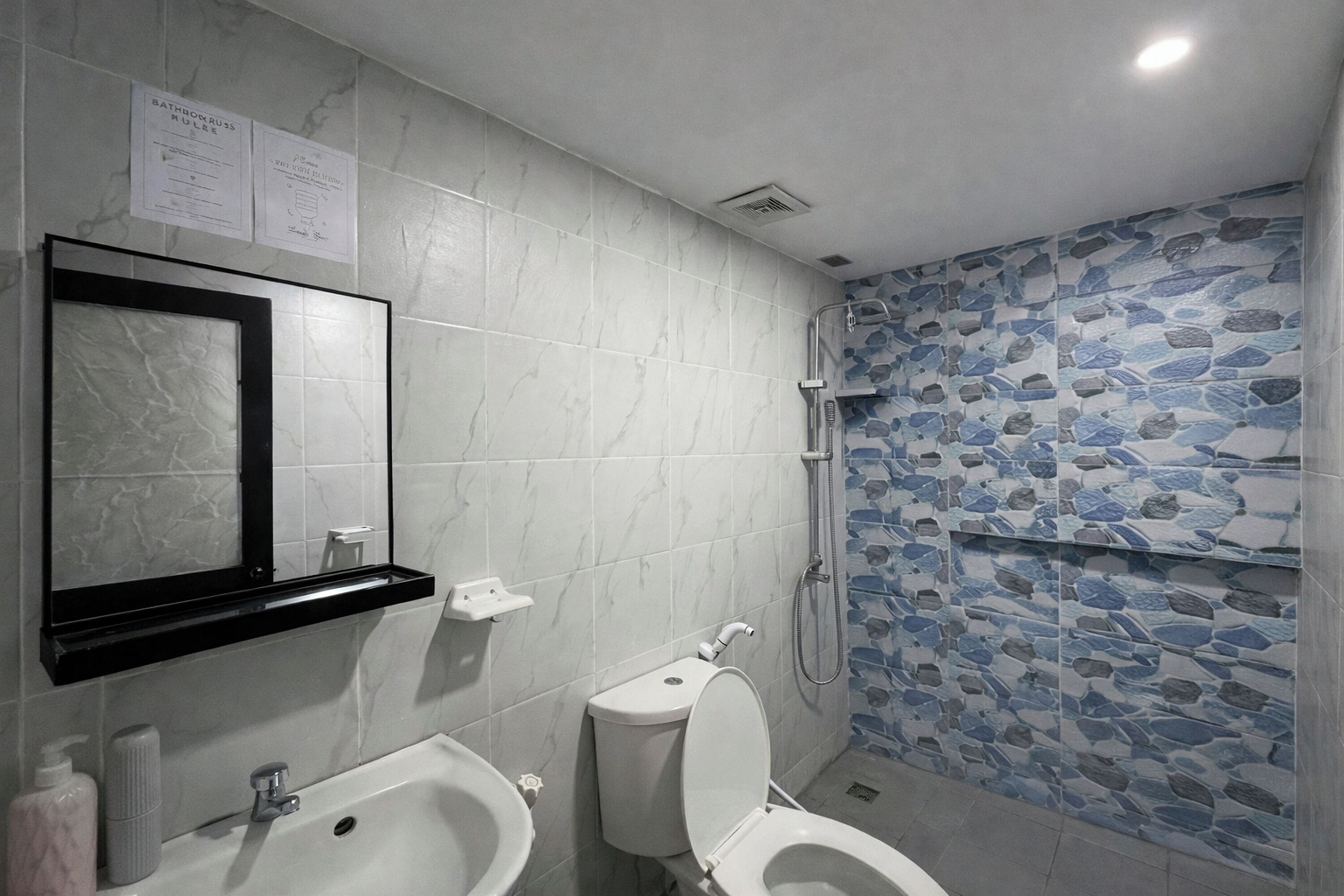Une salle de bain avec un lavabo en marbre, des toilettes, un miroir et une douche dont le mur est décoré d’un motif imitant la pierre.