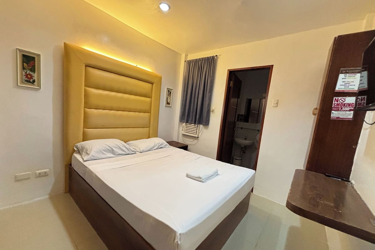 Habitación de hotel con cama, mesita de noche, unidad de aire acondicionado montada en la pared, puerta con espejo y un cartel de 'No Fumar'.