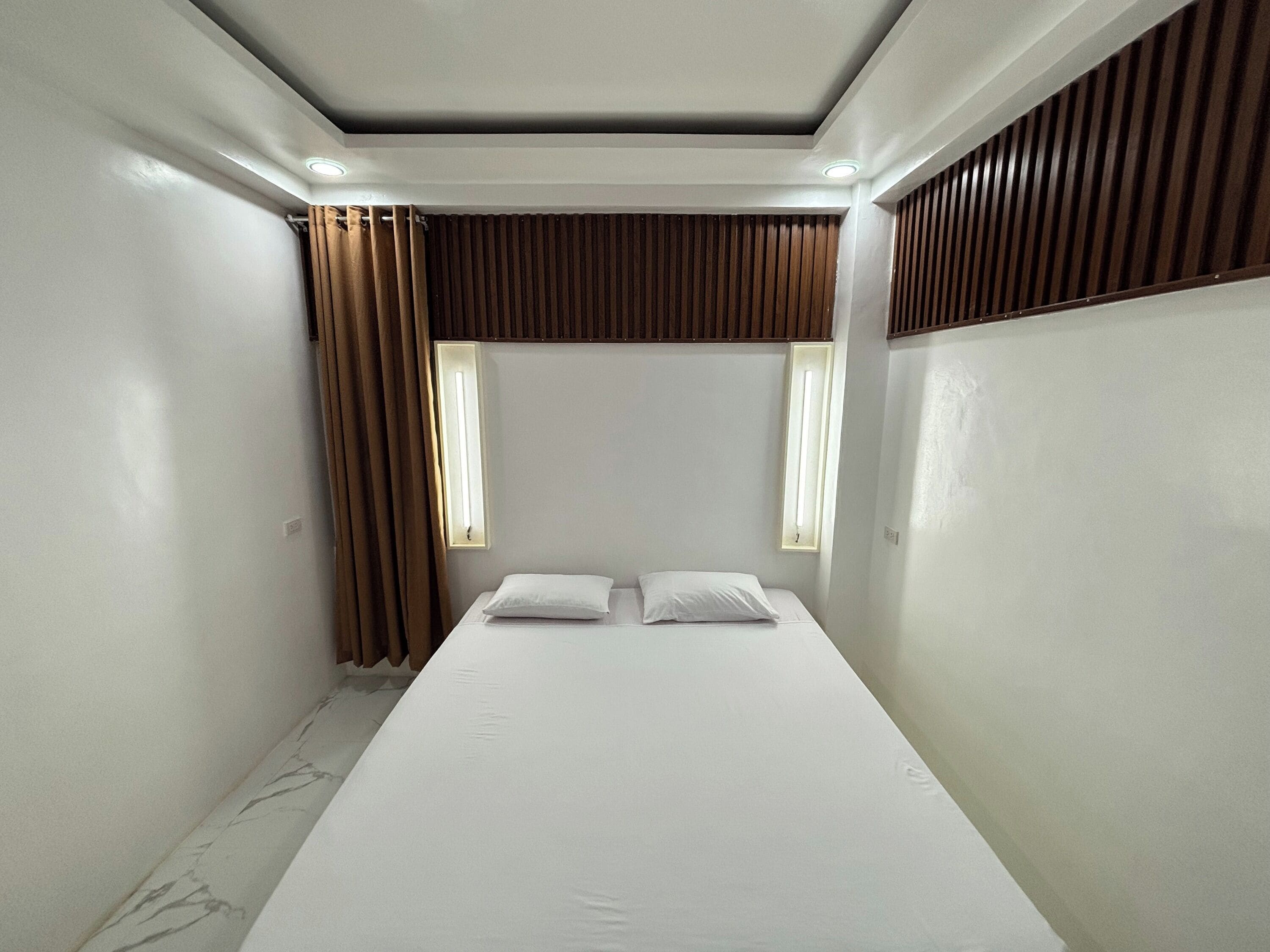 Ein kleines, modernes Hotelzimmer mit einem Einzelbett, zwei Fenstern und einer Decke mit eingelassener Beleuchtung.