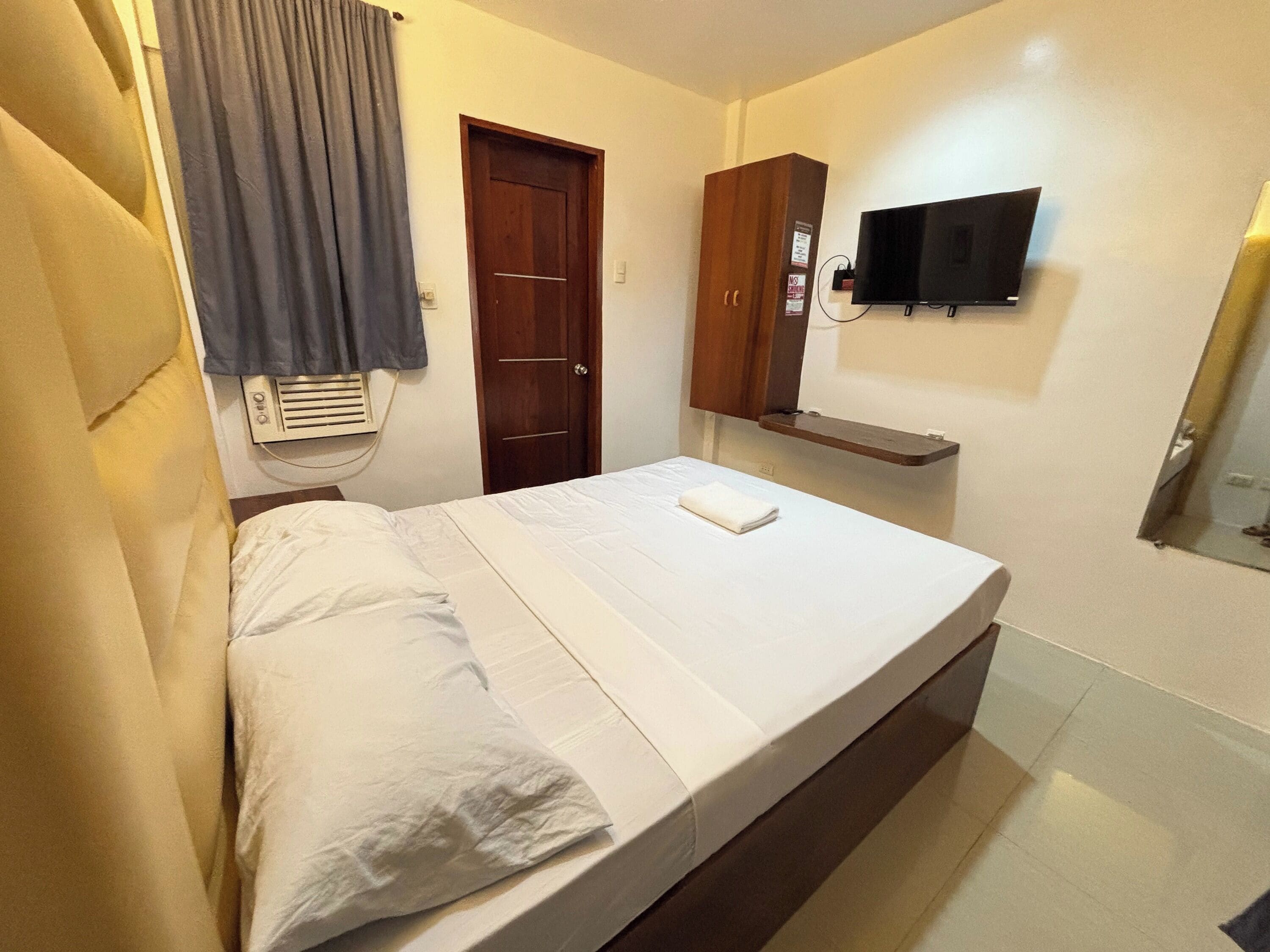 A single bed with white linens, a wooden wardrobe, a mounted television, and a small shelf.