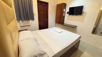 A single bed with white linens, a wooden wardrobe, a mounted television, and a small shelf.