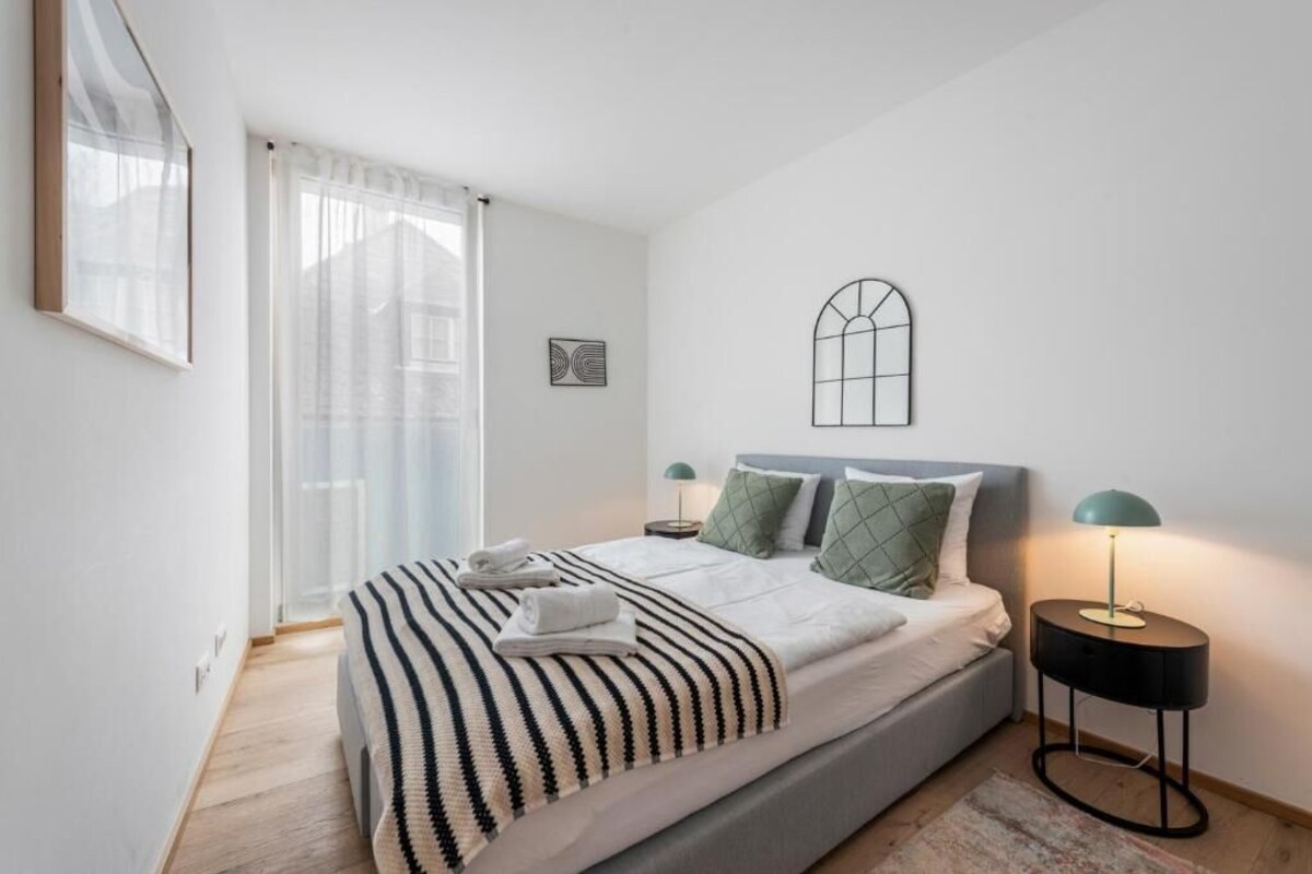 A modern bedroom with a bed, bedside lamps, a window with curtains, and a wall-mounted picture.