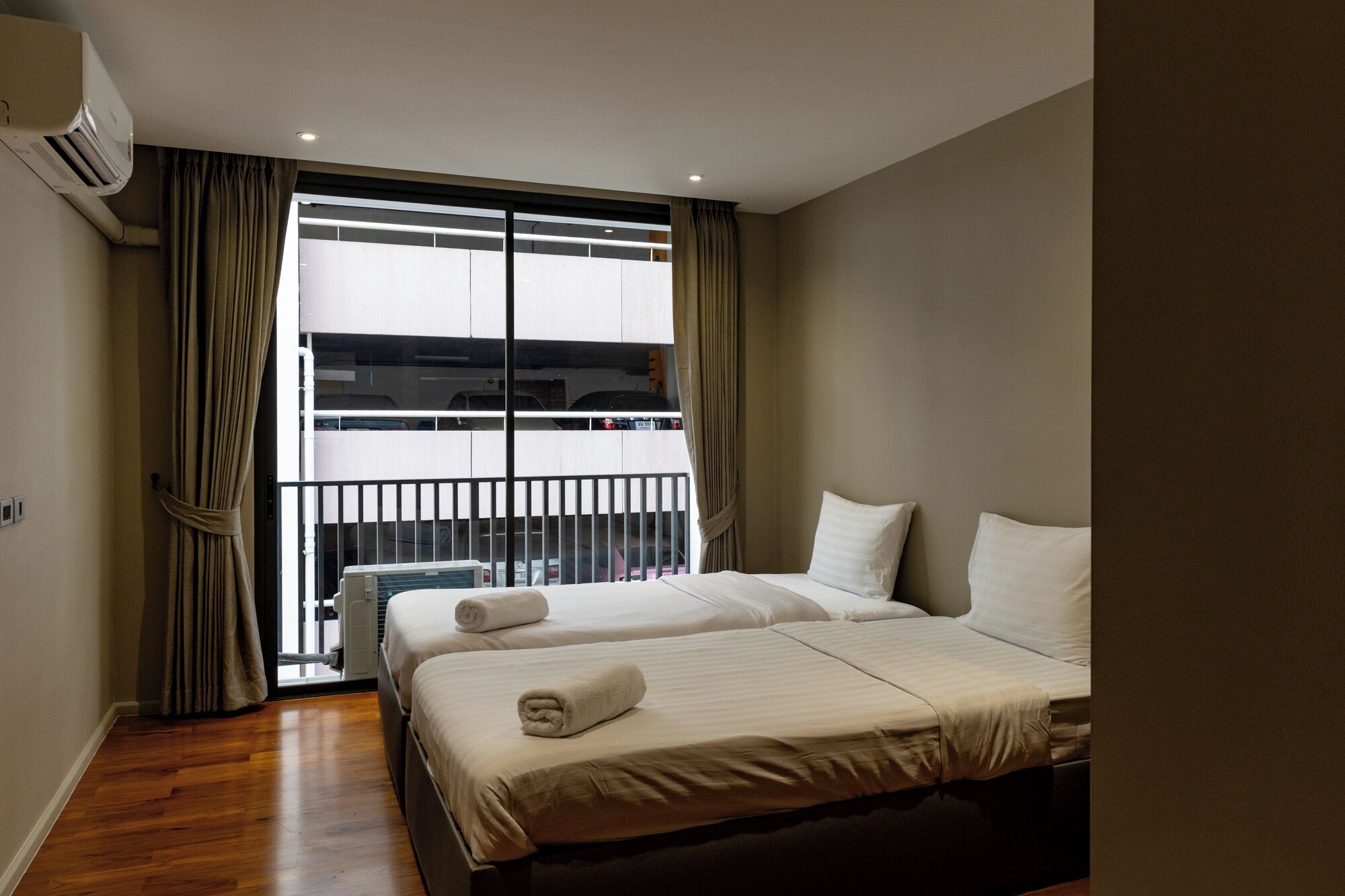 A hotel room with two beds, an air conditioning unit, a window with curtains, and a view of a parking garage.