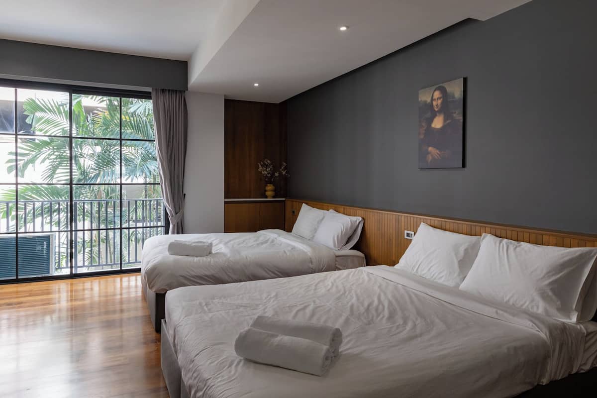 A hotel room with two beds, a large window with a view of palm trees, a painting on the wall, and a wooden headboard.