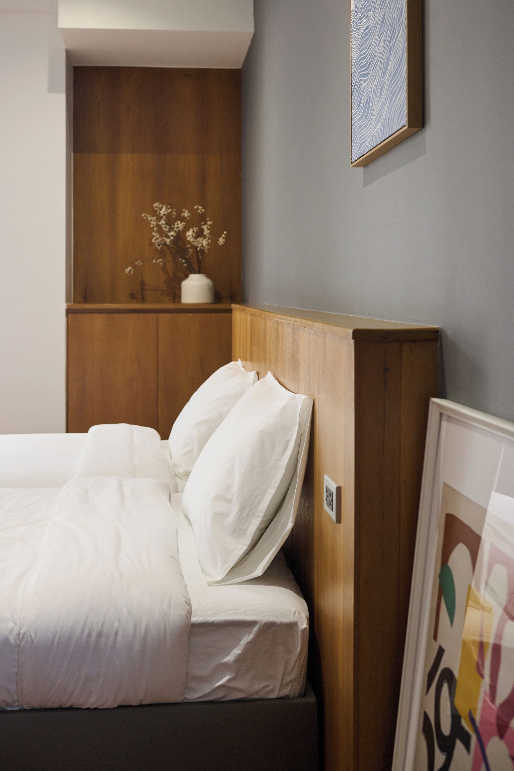 A bed with white bedding, a wooden headboard, and a small white vase with flowers on a wooden shelf.