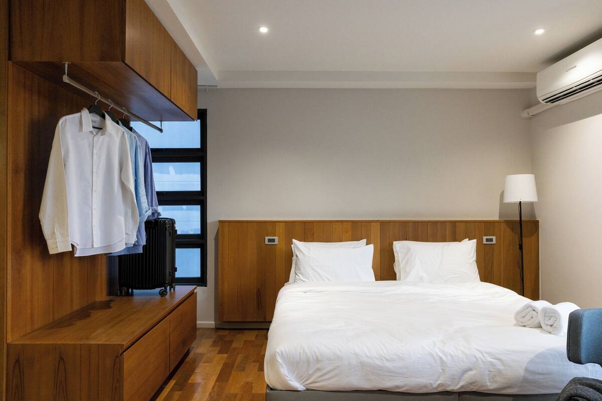 A hotel room with a wooden wardrobe, a bed with white linens, a bedside lamp, and a view through the window.