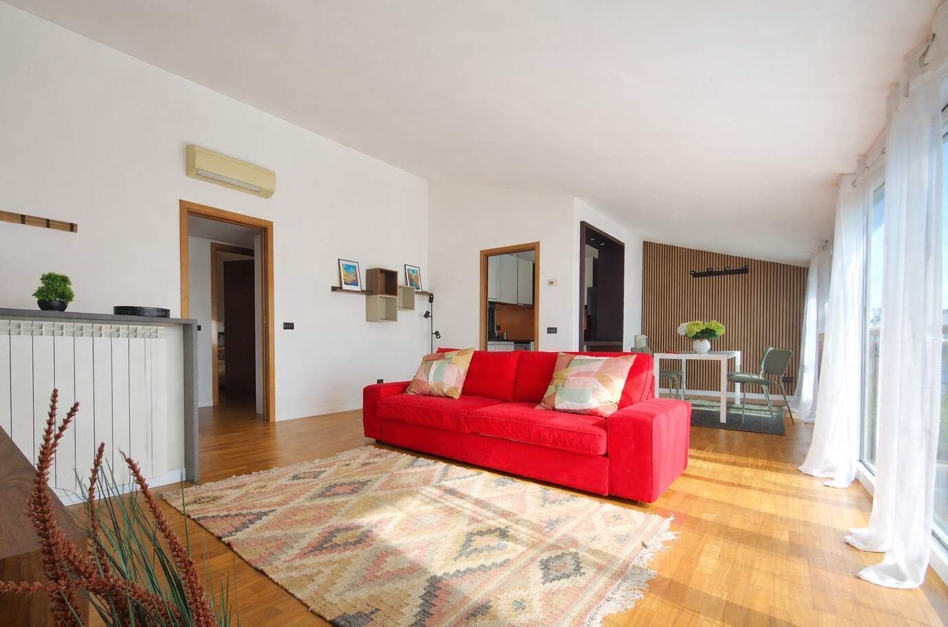 Een woonkamer met een rode bank, een kamerbreed tapijt en een eethoek met tafel en stoelen.