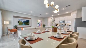 A modern dining area with a long table set for six, a sofa, a TV, and a kitchen in the background. - Luxury home in the best location (Four Corners)