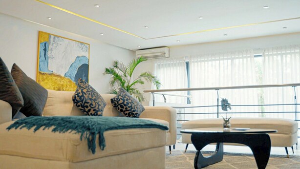 A modern living room with a beige sofa, patterned cushions, a blue throw, and a black coffee table. There is a large abstract painting on the wall, a potted plant, and a window with sheer curtains. - The Mews by Stay Series (Lagos)