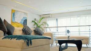 Ein modernes Wohnzimmer mit einem beigefarbenen Sofa, gemusterten Kissen, einer blauen Decke und einem schwarzen Couchtisch. An der Wand hängt ein großes abstraktes Gemälde, eine Zimmerpflanze steht dort, und es gibt ein Fenster mit durchsichtigen Vorhängen.