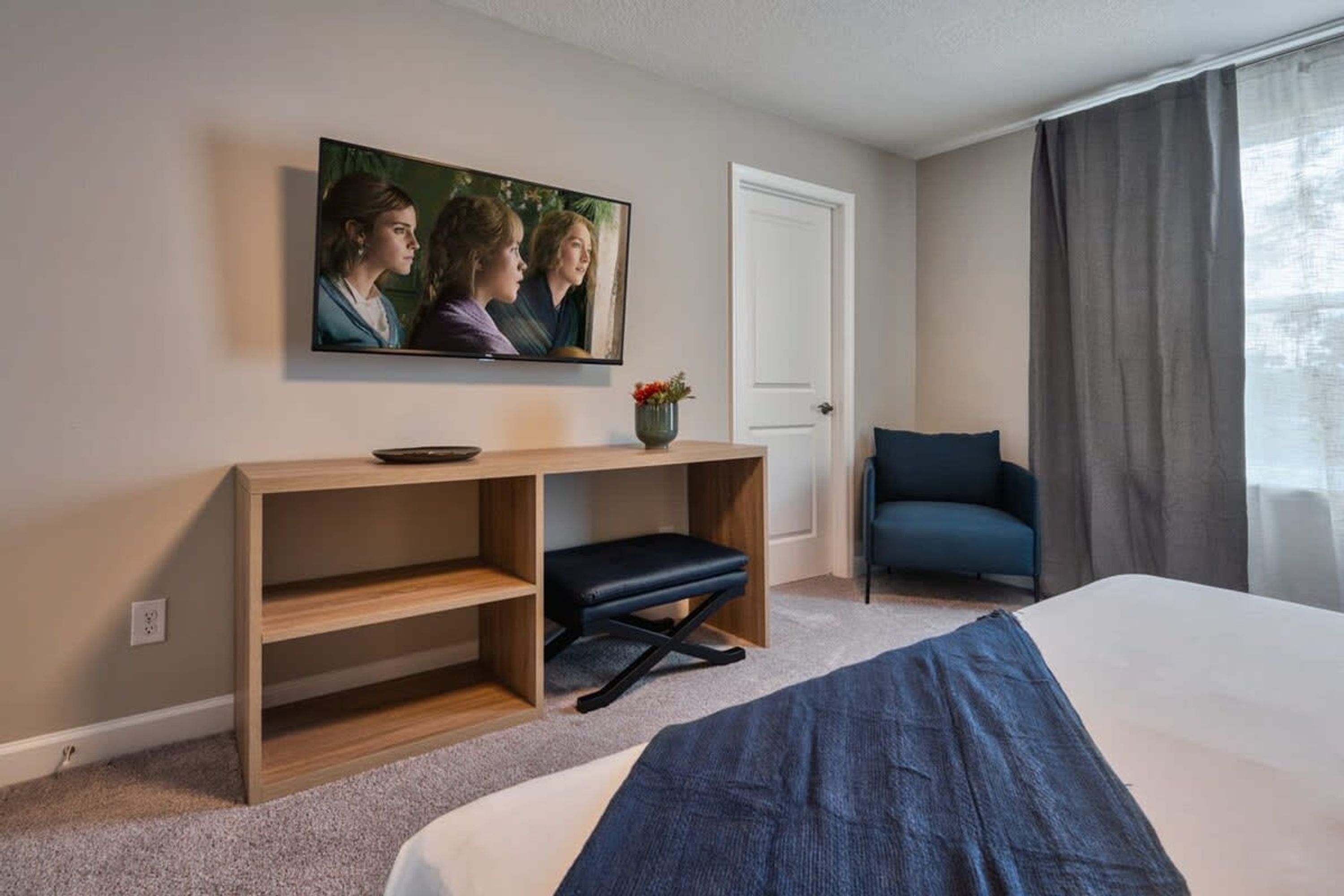 Et hotelværelse med seng, skrivebord med stol, fjernsyn og et vindue med gardiner.