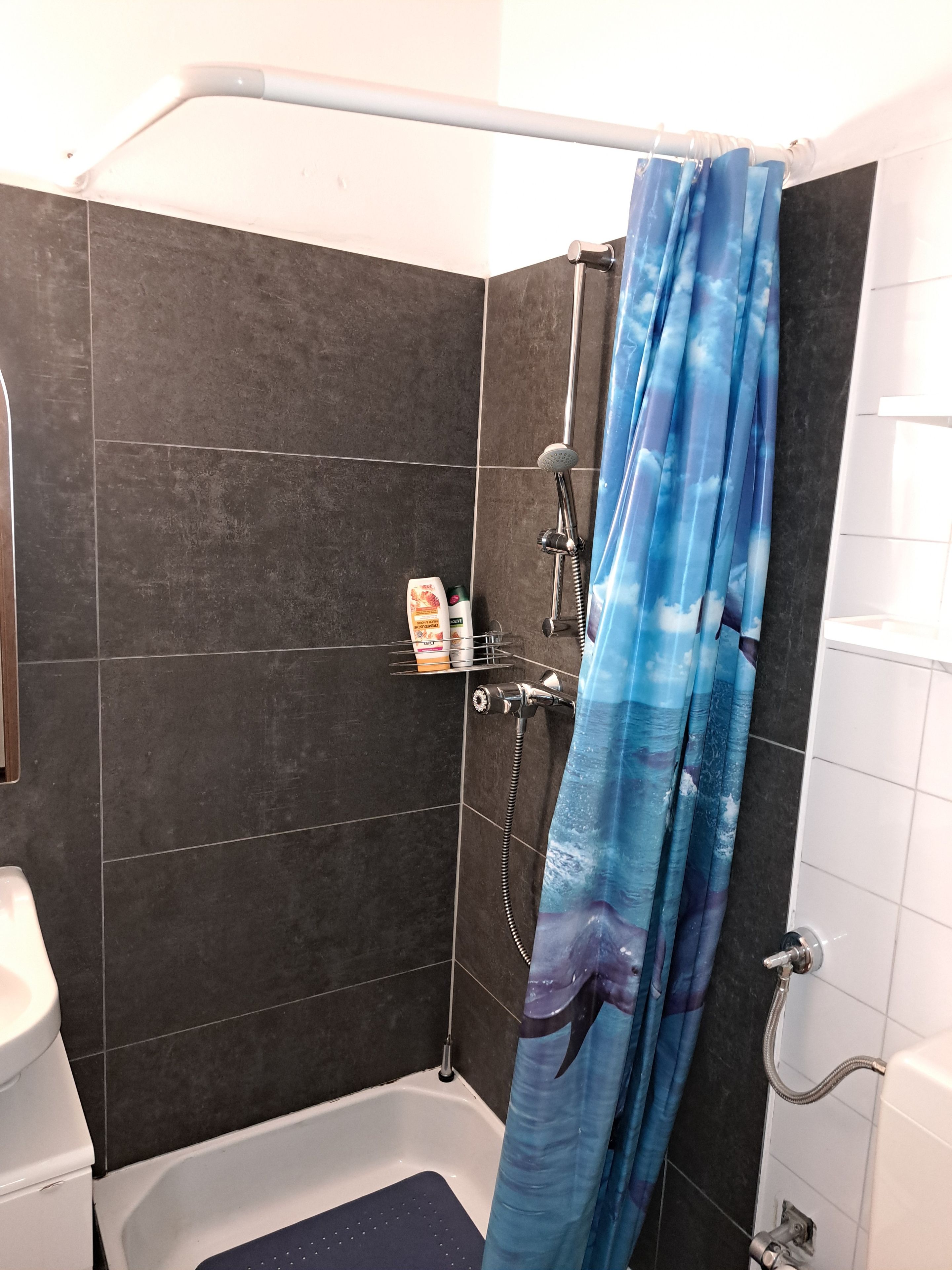 Zona de ducha con pared de azulejos oscuros, cabezal de ducha, ducha de mano y cortina de baño azul con diseño de olas.
