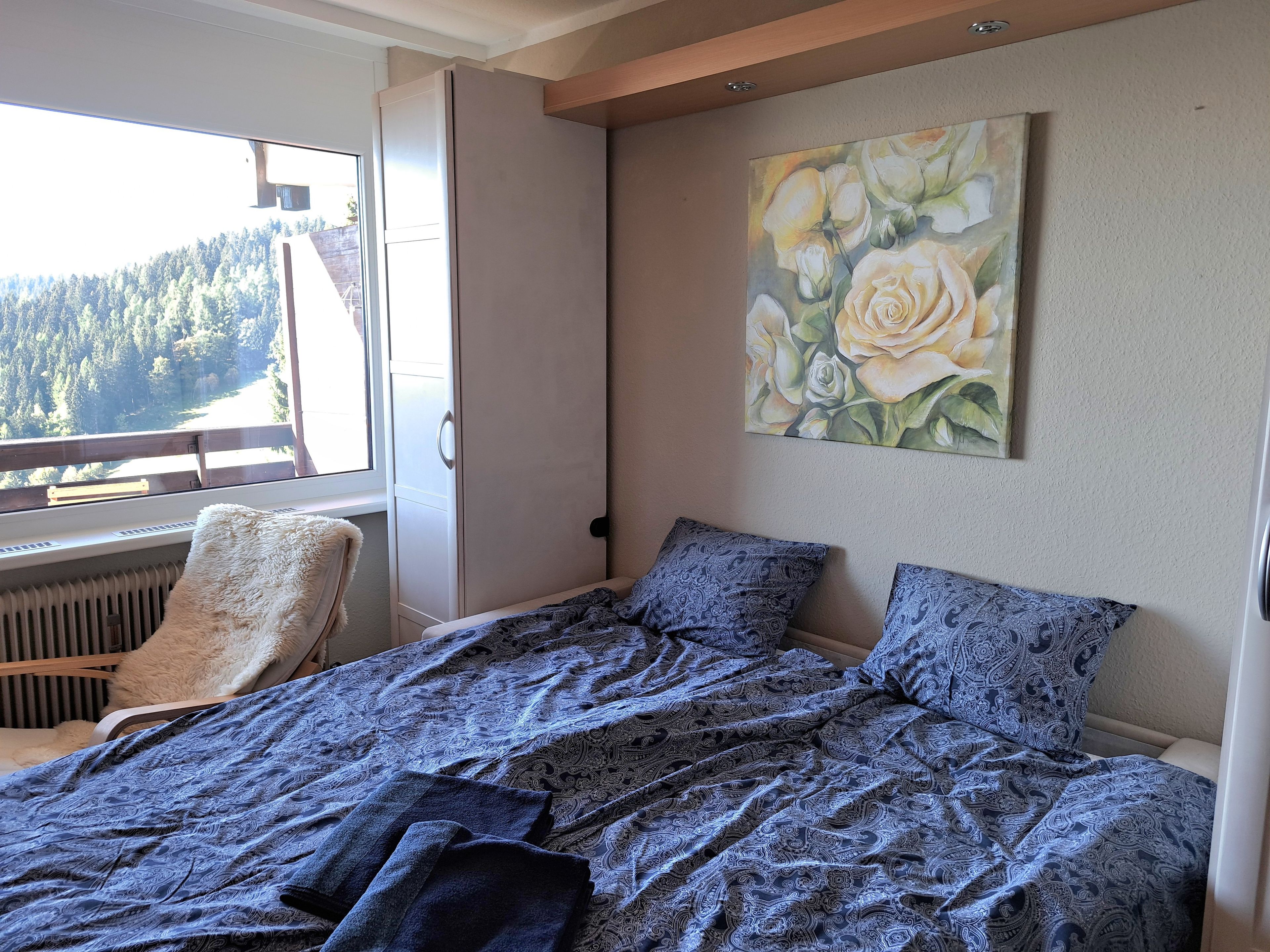 A bedroom with a bed, a chair, a radiator, a window with a view of trees, and a painting of flowers on the wall.