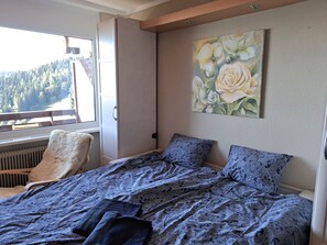 Quarto com cama, uma cadeira, radiador, janela com vista para árvores e um quadro de flores na parede.