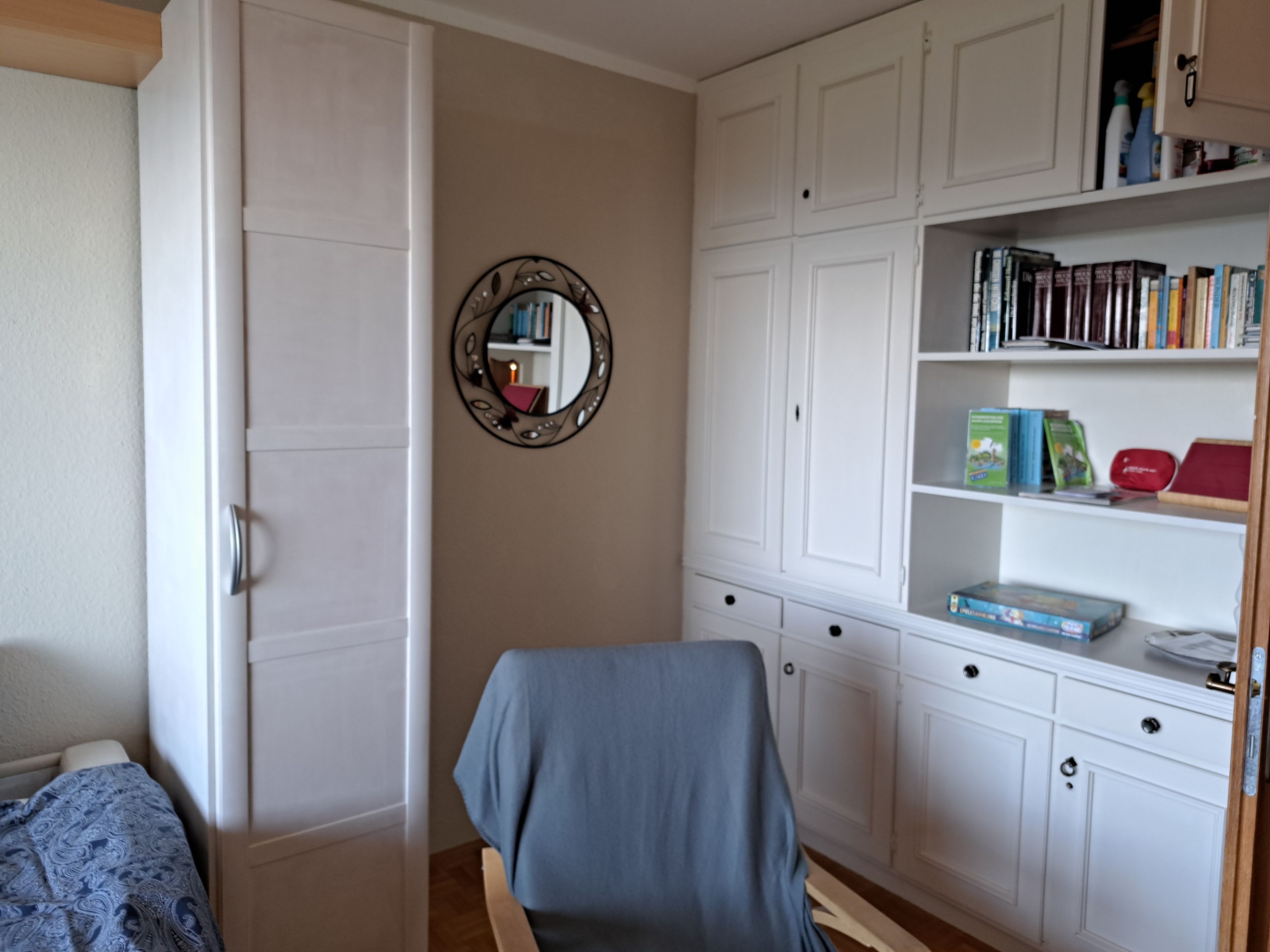 A room with a bed, a chair, a bookshelf, and a mirror.