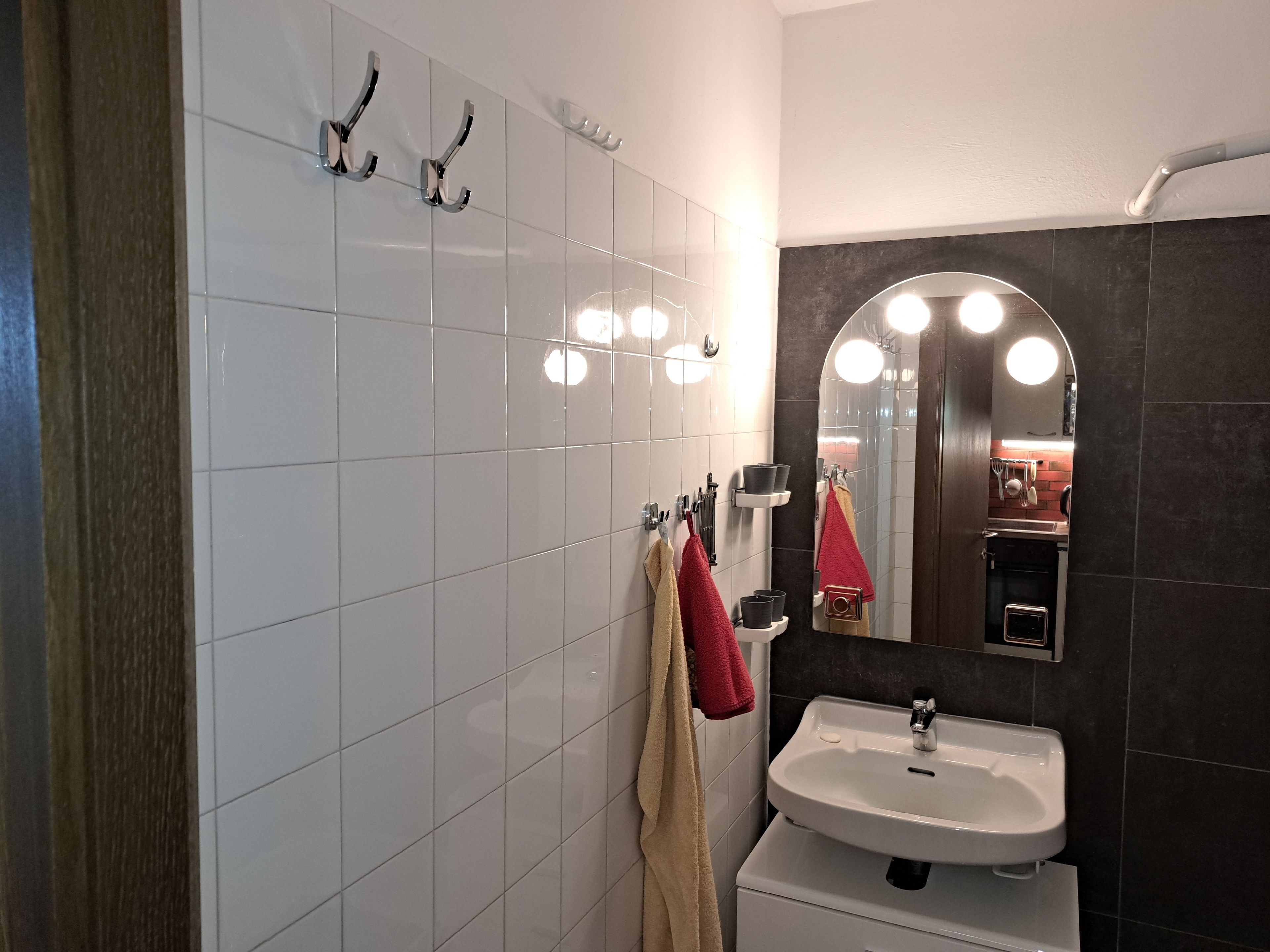 Une salle de bain avec un mur recouvert de carreaux blancs, un lavabo, un miroir avec éclairage intégré et des serviettes.