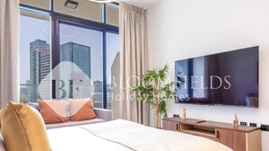 A modern hotel room with a large bed, a flat-screen TV, a wooden console, and a view of the city skyline. - Bloomfileds Executive Studio Al MA (Abu Dhabi)