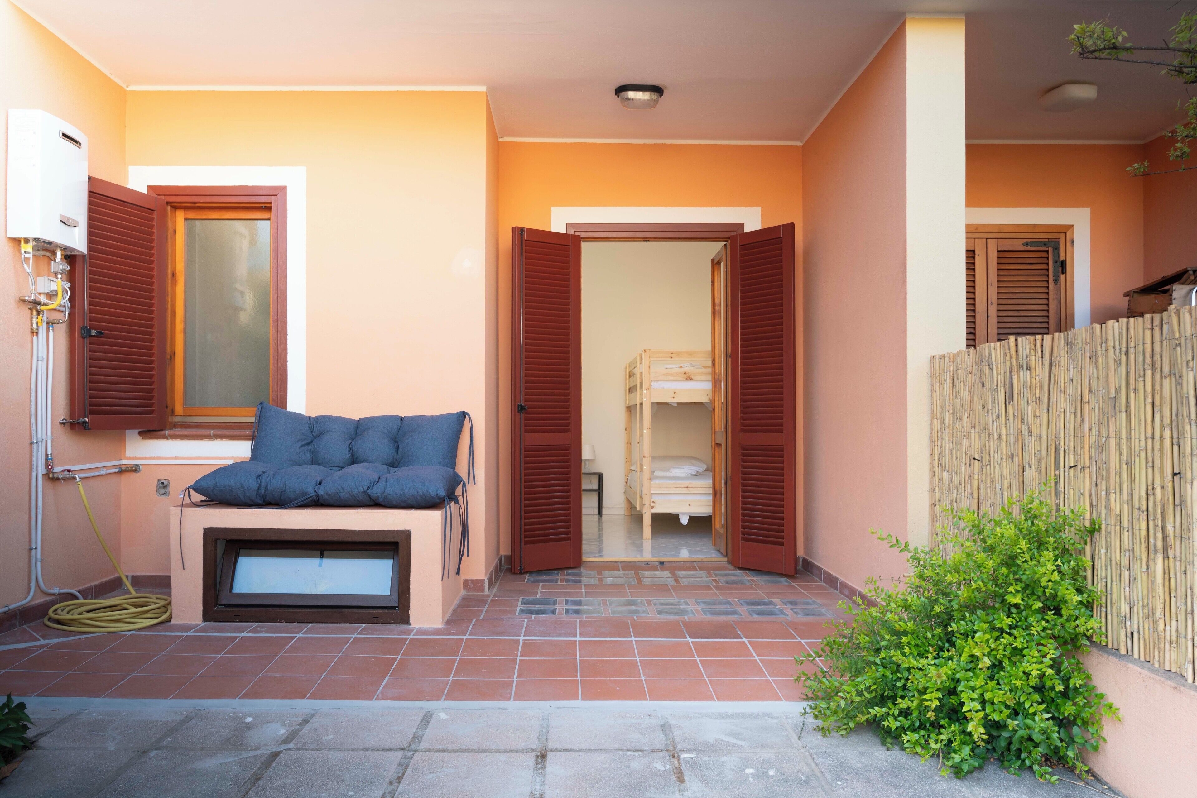A room with a bed, a window with shutters, a small table with a cushion, and a wooden fence outside.