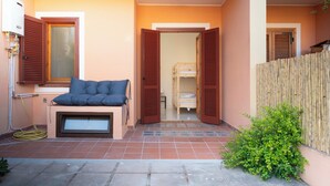 Quarto com cama, janela com persianas, pequena mesa com almofada e cerca de madeira lá fora.