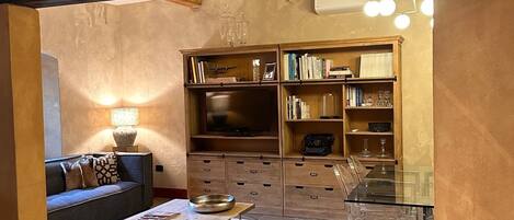Ein geräumiges Wohnzimmer mit einem durchbrochenen Holzboden, einem dunklen Sofa, einem Couchtisch und einem Glastisch. Ein Bücherregal voller Bücher und Dekorationselementen sowie ein Fernseher auf einem Holzgestell sind ebenfalls vorhanden.