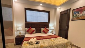 A bedroom with a large bed, two bedside tables, a window with a grid pattern, a door, and a wall-mounted painting. - Fortune Home Service Apartment 3Bhk J150 (New Delhi)
