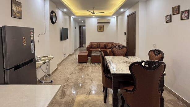 A modern living space with a dining area, a sofa, a TV, and a ceiling fan. - Fortune Home Service Apartment 3Bhk J150 (New Delhi)