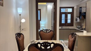 A dining area with a marble table and brown upholstered chairs, a ceiling light, and a kitchen with cabinets and a microwave. - Fortune Home Service Apartment 3Bhk J150 (New Delhi)