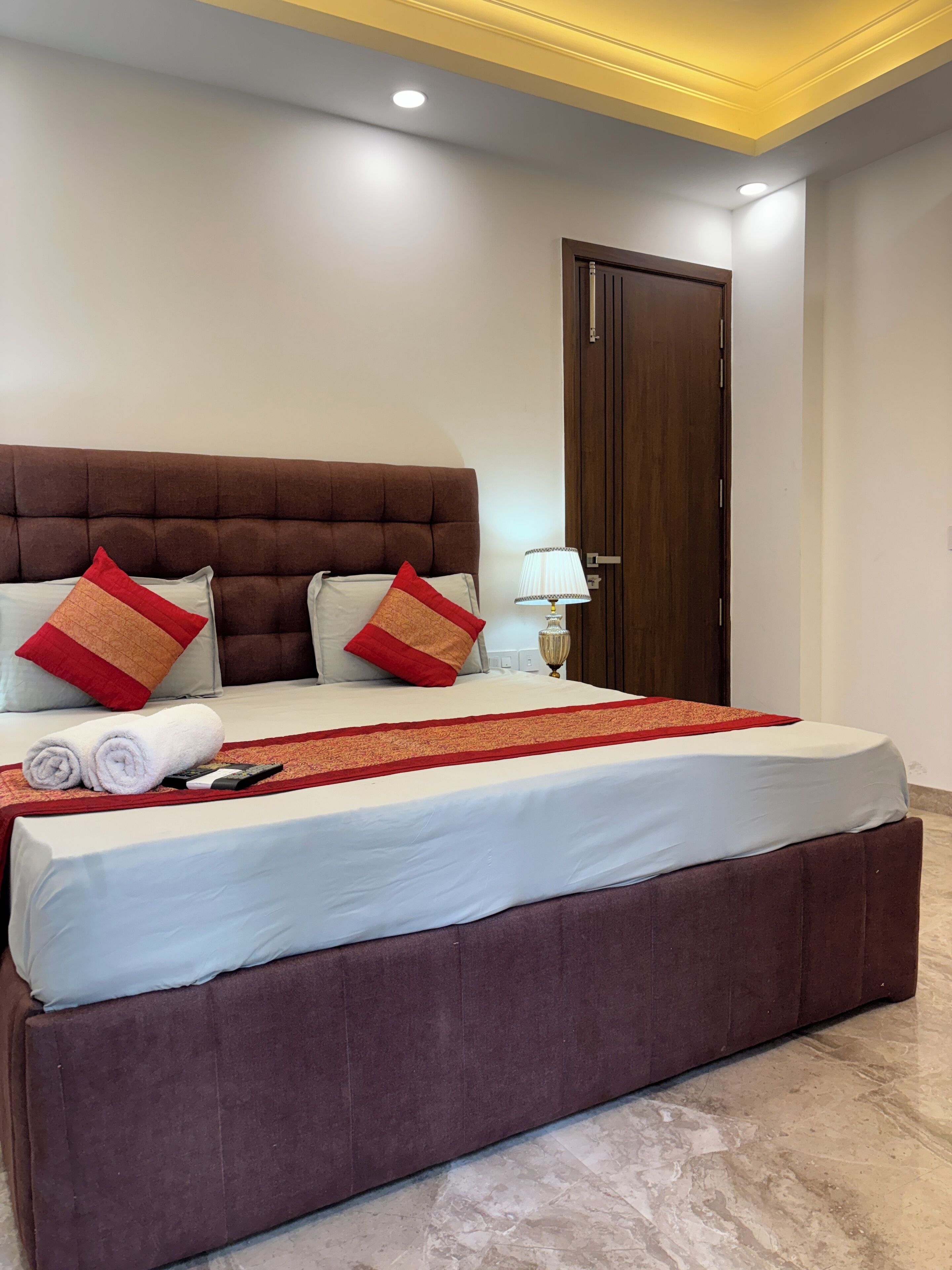 A hotel room with a large bed, a wooden door, a nightstand with a lamp, and a marble floor.