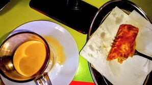 A table with a cup of coffee, a smartphone, and a plate with a piece of food. - Flamingo Hotel Bombolulu  (Mombasa)