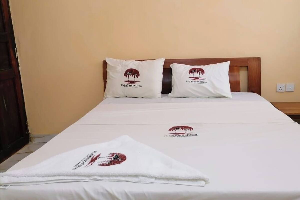 Egy gondosan megterített ágy fehér ágyneművel és két párnával, melyeken a Flamingo Hotel logója látható. Ugyanaz a logó díszíti a fehér törölközőt is, amely az ágyon van.