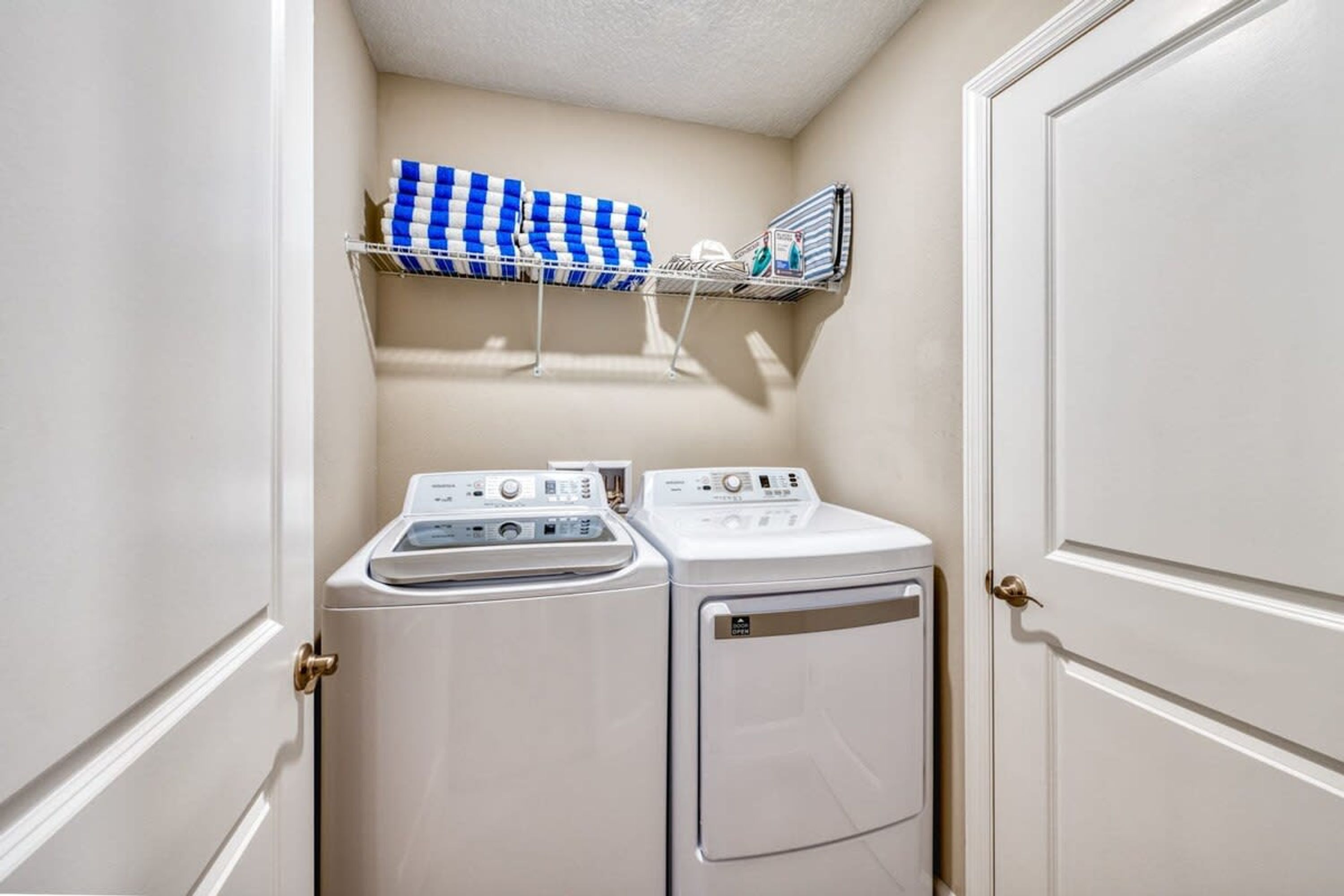 A laundry room with two stacked washers and dryers, a metal shelf with folded towels, and a white door.