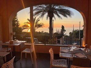 An outdoor dining area with tables and chairs, a view of the sunset over the sea, and a palm tree in the background. - UNICO Cannamele Escape Tropea (Parghelia)