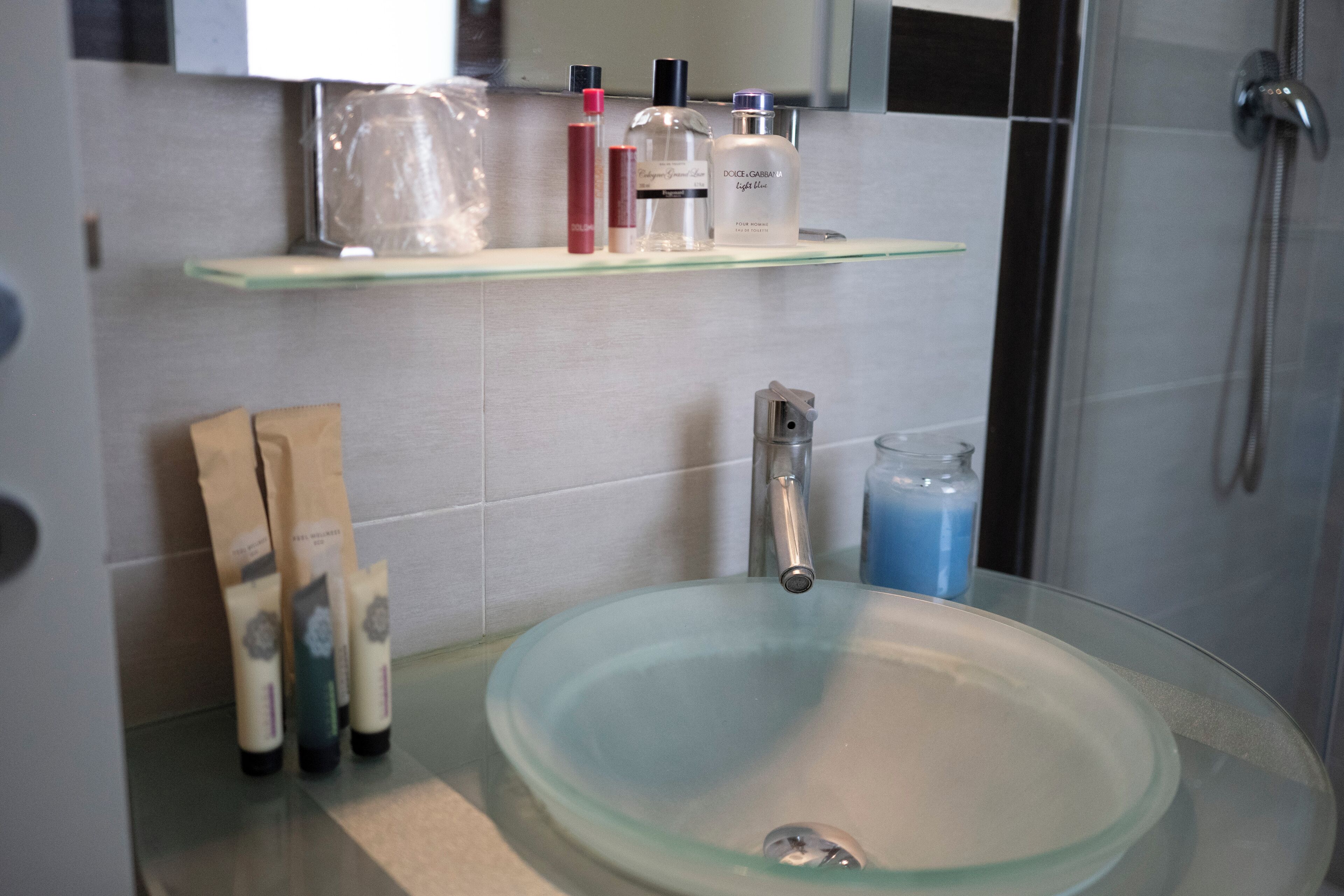 Un baño con lavamanos de vidrio, ducha y repisa con artículos de aseo.