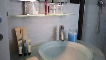 Un baño con lavamanos de vidrio, ducha y repisa con artículos de aseo.