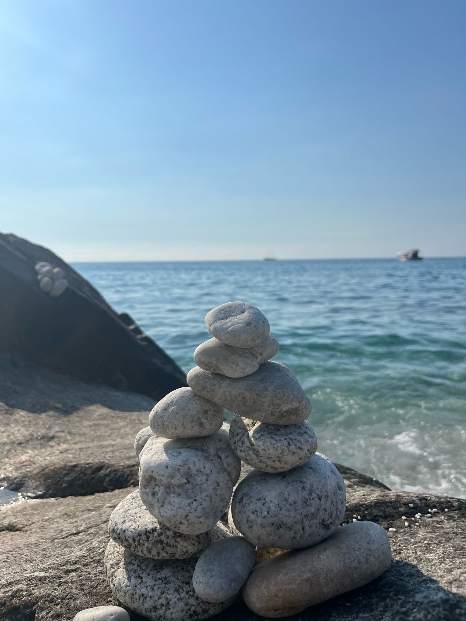 Ein Stapel glatter, abgerundeter Steine an einer felsigen Küste mit ruhigem Wasser und einem Boot im Hintergrund.