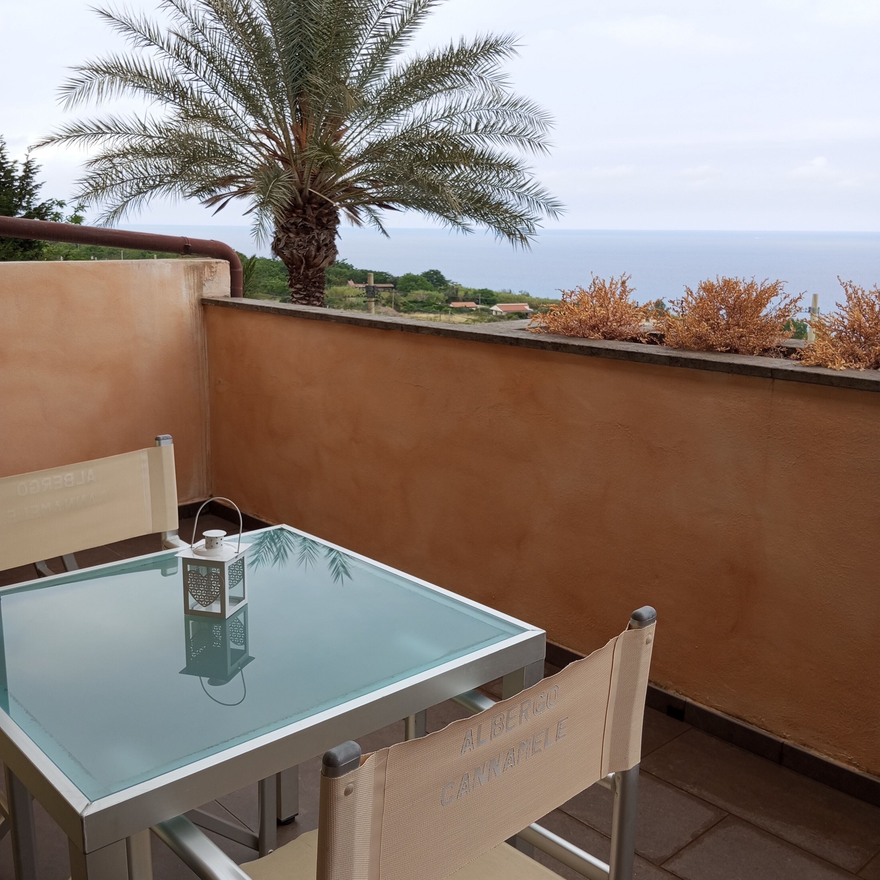 Een glazen tafel met een stoel, een waterfles en een kleine lamp op een balkon met uitzicht op water.