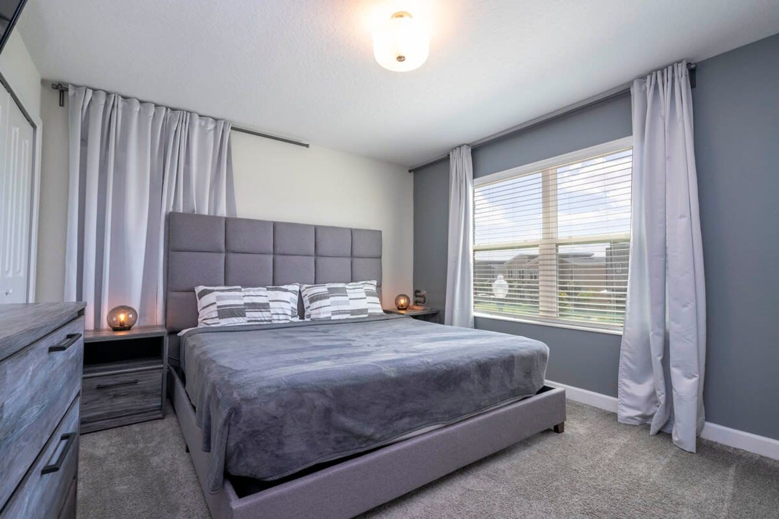 A modern bedroom with a large bed, grey bedding, a headboard, bedside tables, and a window with curtains.